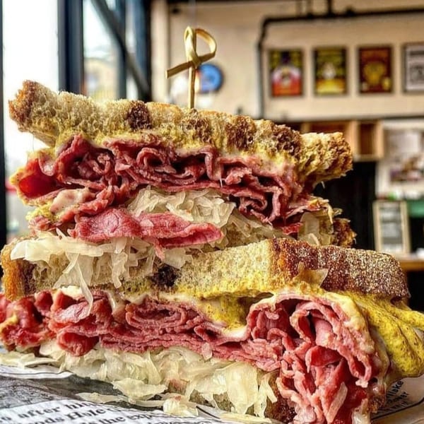 A towering pastrami and sauerkraut sandwich on rye bread with mustard, held by a decorative toothpick.